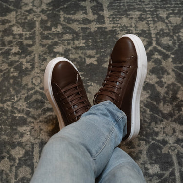 Lifestyle image of a person wearing brown leather Sneakers by Shoescoo, crossed-leg view on a patterned rug.